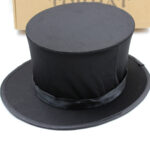 Chapeau haut de forme noir posé sur un fond blanc avec un carton marron à l'arrière avec des inscriptions noires dessus.