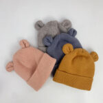 Bonnets pour bébé fille avec des oreilles d'ourson. Un bonnet rose,un camel, un bleu et un gris posés sur une table blanche.