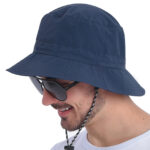 Homme souriant portant sur sa tête un chapeau bleu et des lunettes de soleil. L'homme porte également un t-shirt blanc et est positionné de profil.