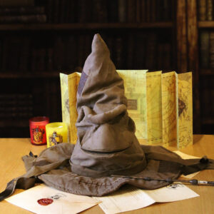 Le choixpeau magique issu de la sage Harry Potter est posé sur une table et sourit. Une carte et des bougies se trouvent en arrière plan. Le choixpeau magique est au dessus d'une lettre et d'une enveloppe de Poudlard ainsi que d'une baguette magique.