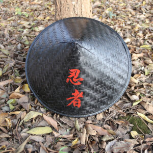Chapeau de samouraï noir posé contre arbre et sur un sol couvert de feuilles mortes marrons et vertes.
