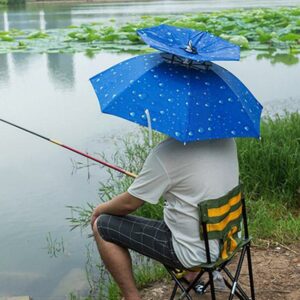 Homme qui pêche avec un chapeau parapluie bleu à motif goutte de pluie sur la tête. L'homme est assis sur un chaise et fait face à une rivière. Il porte une cane à pêche.