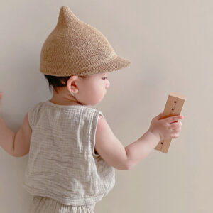 Bébé de dos avec un chapeau en paille pointu sur la tête et portant un bout de bois dans la main droite.