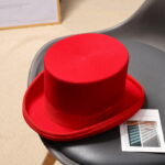 Chapeau haut de forme rouge en feutre posé sur une table basse ronde grise à côté d'un flyer. Derrière se trouve une chaise grise et le bord d'un tableau posé par terre.