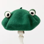 Béret vert en laine avec des yeux de grenouille sur le dessus. Le béret et posé sur une barre en bois blanche.