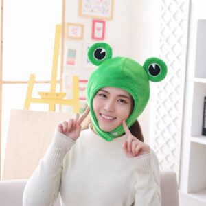 Femme avec un pull blanc portant un chapeau vert avec des yeux de grenouilles sur le dessus. Derrière se trouve des meubles d'une chambre avec un chevalet de peintre