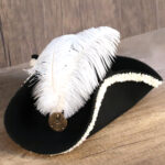 Chapeau tricorne noir avec une grosse plus blanche sur le dessus et un médaillon dorée sur le devant. Bordure du chapeau blanches.