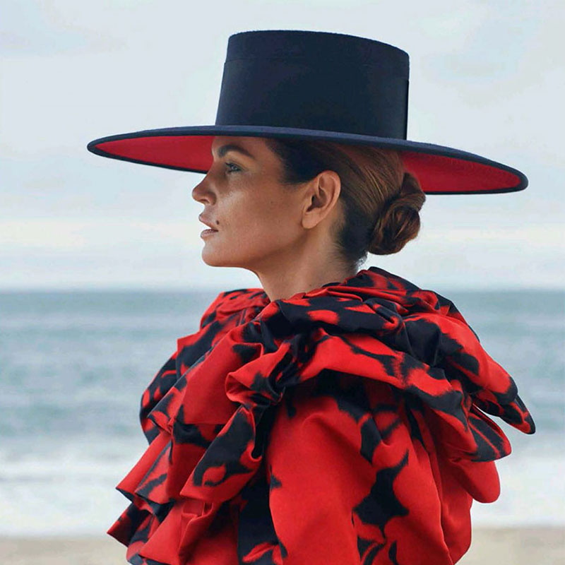 Jolie femme qui porte une tenue typiquement espagnole avec des froufrous rouges et noirs. Elle est sur une plage et porte un chapeau.