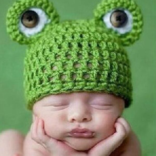 Tête de bébé avec les mains posées sur le menton portant un bonnet en laine vert avec des yeux de grenouille sur le dessus.
