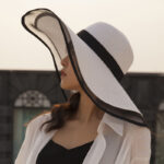 Chapeau capeline, à larges bords, en paille blanche, avec bordures en tissu organza noir porté par une femme.